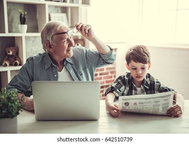 Grandpa And Grandson Resting At Home. Handsome Old Man Is Using A Laptop While His Boy Is Reading A Newspaper