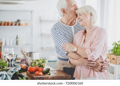 Grandpa Hugs And Kisses Smiling Grandma During Cooking Healthy Meal In Kitchen