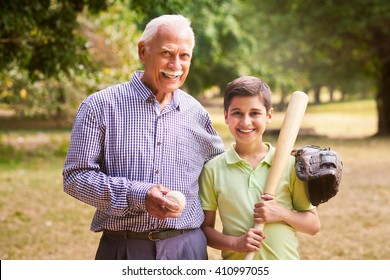 Grandpa Spending Time With Grandson: Portrait Of Senior Man Playing Baseball With His Grandchild In Park. The Old Man Embraces The Young Kid Holding The Bat, Smiling And Looking At Camera