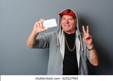 Grandpa Takes A Selfie. Studio Shot Of An Old Funny Man Who Takes A Photo Of Himself With A Peace Sign. Grandfather's Style Includes Light Gray Hoodie Over A Black T-shirt, A Gold Chain And A Red Cap.