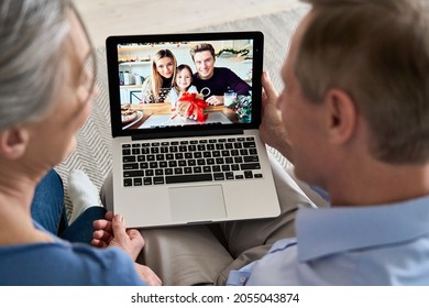 Grandparents Couple Holding Computer Video Calling Family With Grandkid On Laptop Screen Having Fun Enjoying Talking Online, Greeting With Gift During Virtual Distant Meeting Chat, Over Shoulder View.