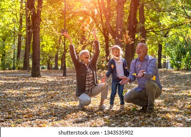 Grandparents And Grandchild Throwing Leaves In Park And Having Fun Together