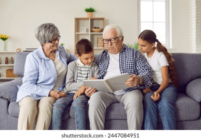 Grandparents and grandchildren reading a book together. Happy grandfather, grandmother and kids sitting on the sofa at home and reading a story from a children's book. Family, leisure concept - Powered by Shutterstock