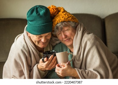 Grandparents in knitted caps wrapped in warm blanket hold cups of hot tea or coffee, feeling cold at home. Happy couple. - Powered by Shutterstock