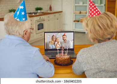 Grandparents In Party Hats Sitting At Laptop, Video Calling Their Grandchild And Wishing Him Happy Birthday. Family With Little Child Having Virtual Celebration At Home Due To Lockdown And Quarantine