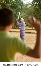 Grandparents Spending Time With Grandson: Senior Man Playing Baseball With His Grandson In Park. The Old Man Holds The Bat, While The Kid Prepares To Throw The Ball