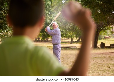 Grandparents Spending Time With Grandson: Senior Man Playing Baseball With His Grandson In Park. The Old Man Holds The Bat, While The Kid Prepares To Throw The Ball