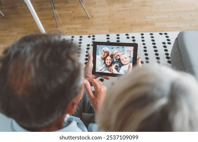 Grandparents talking to their family and grandchildren over a video call. Senior couple sitting on the couch in the living room, talking to family over video call on digital tablet. - Powered by Shutterstock