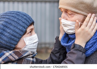 Grandson Grandchild Puts A Protective Mask On His Elderly Grandmother Face Outdoors. Danger Of Infection With Coronavirus Covid-19. Family Care. Quarantine Caring For The Elderly Relatives