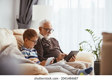 Grandson And Grandfather Using Digital Tablets And Surfing The Net While Relaxing On The Sofa.