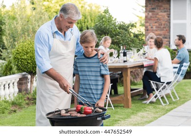 Grandson Helping Grandfather In Barbeque. Happy Grandpa Teaching Grandson How To Cook Food. Grandson Enjoying Watching Grandfather Cooking Barbeque.