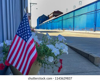 Granite City, IL—July 27, 2018; American Flag In Pot Of Red And White Flowers With Blue Buildings Of United States Steel Granite City Mill Across The Road After Grand Reopening Of Furnace.