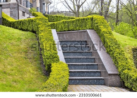 The granite steps of the garden stairs on the slope of the park, framed on both sides by trimmed bushes, lead to the facade of the house in the background.