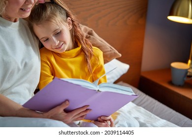 Granny And Child Girl Read A Book Before Bed. Evening Ritual Before Bedtime, Communication Between Generations, Retired Female Is Spending Time With Grandchildren, Relaxing And Teaching