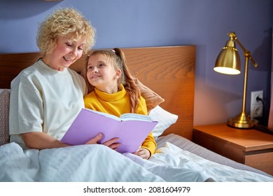 Granny And Grandchild Read A Book Before Bed. Evening Ritual Before Bedtime, Communication Between Generations, Happy Female Is Spending Time With Grandchildren, Relaxing And Teaching