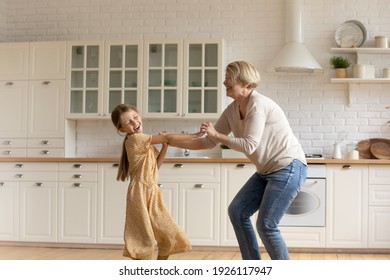 Granny teaches to dance. Laughing little girl mature aged grandmother hold hands move by energetic music having fun. Active senior grandma entertaining small grandchild kid sing jump at home kitchen - Powered by Shutterstock