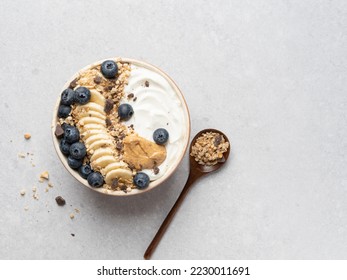 Granola bowl. Yogurt and muesli with fresh banana slices, blueberry and peanut butter in ceramic bowl. Top view. Copy space. Gray background. Healthy vegetarian breakfast or lunch. Morning tasty meal. - Shutterstock ID 2230011691