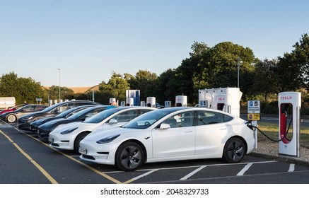 Grantham, Lincolnshire, England -
September 26 2021: Line Of Tesla Electric Car Chargers With Cars On Charge At A Motorway Service Area  On A Clear Autumn Evening In Warm Light From Setting Sun.