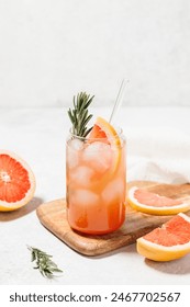 Grapefruit cocktail with rosemary garnish in a glass, served on a wooden cutting board. Vibrant, refreshing drink perfect for summer menu or cocktail recipes. Greyhound, lemonade, paloma, mocktail