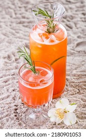 Grapefruit Lemonade With Rosemary In A Tall Glass Decanter With Lime Wedges And Ice Inside. Nearby Is A Stemmed Glass With Lemonade And A Sprig Of Ozmarine. The Crockery Stands On A Sandy Backdrop, Ne