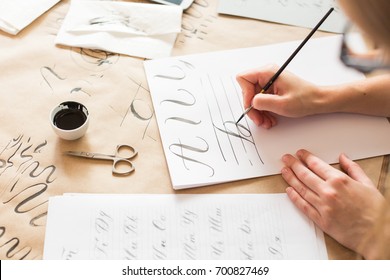 graphic design, handwriting, creation concept. tender little hands of female painter inscribing ornamental decorated letters on the white lined paper with help of thin tip of the brush and black ink