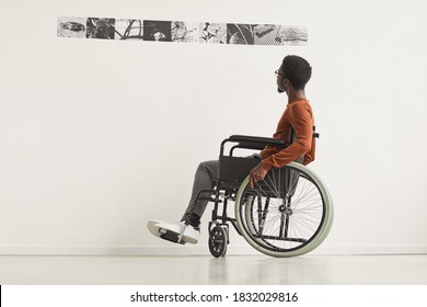 Graphic full length portrait of young African-American man using wheelchair and looking at paintings while exploring modern art gallery exhibition, copy space