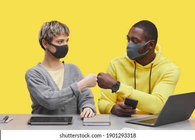 Graphic portrait of two modern business people wearing masks and bumping fists while working together at desk against pop yellow background, copy space