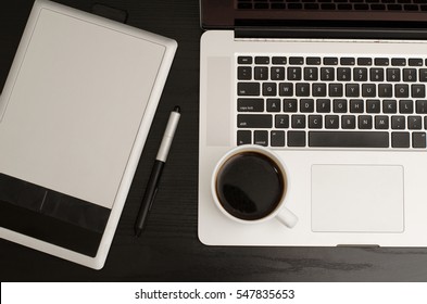 Graphics tablet with a pencil, part of the notebook and cup of coffee on a black wooden table, close up