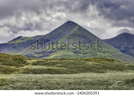 A grass covered rugged mountain with with a grey bare rock pointed conical peak with short grassland in the foreground