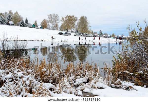 The grass on the snowy Islands in the water near the\
shore autumn pond with bushes in the foreground and people anglers\
on the opposite Bank with cars and winter, small colorful cottages.\
Soft focus. 