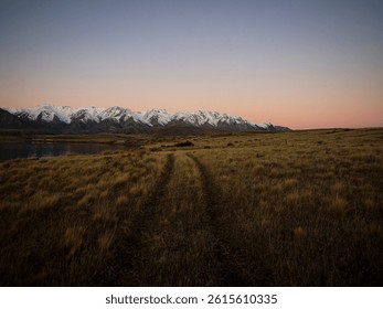 Grass road tire prints leading to idyllic remote calm serene golden hour snow capped Southern Alps mountains, Lake Heron winter sunset landscape, Ashburton Lakes Canterbury South Island New Zealand - Powered by Shutterstock - Get 15% off with code: PIKWIZARD15