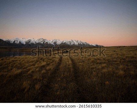 Grass road tire prints leading to idyllic remote calm serene golden hour snow capped Southern Alps mountains, Lake Heron winter sunset landscape, Ashburton Lakes Canterbury South Island New Zealand
