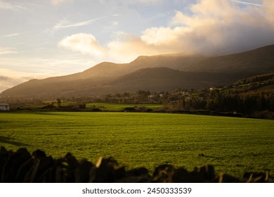A grassy field with a wooden fence in the foreground, under a clear sky cloudy Mourne mountains in Northern Ireland tallest hill sunrise sunlight pouring over ocean sea green lawn stone wall houses - Powered by Shutterstock