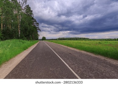 Grassy roadside under heavy, brooding clouds. Asphalt path leads straight ahead into distant greenery