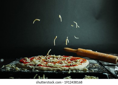 Grated Cheese Floating, Falling Pizza. Pizza Dough With Tomato Sauce And Lots Of Mozzarella. Black Background, Wooden Rolling Pin And Flour.