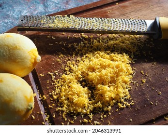 Grated Lemon Zest With Stainless Steel Zester Or Grater Over Wooden Cutting Board On Rustic Blue Table With Fresh Lemons. Process Of Cooking Citrus Fruits, Food Ingredient Preparing, Healthy Eating.