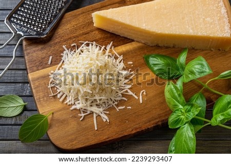 Grated parmesan cheese on a cutting board. Italian grana padano cheese whole and grated, grater and fresh basil herb over wooden background. Delicious hard cheese, dairy product. Top view.
