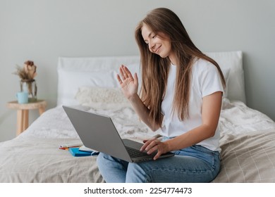 Grateful blonde American young woman in casual sitting on bed with laptop makes video call gesture hello by hand smiles. Student girl with loose long hair talking to parents after relocation to campus - Shutterstock ID 2254774473