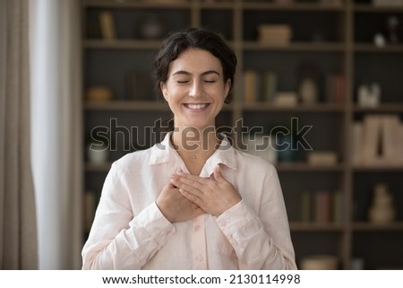 Grateful happy young Hispanic woman with folded hands on chest standing in office. Kind smiling pretty millennial lady with closed eyes feeling thankful, showing appreciation, charity concept.