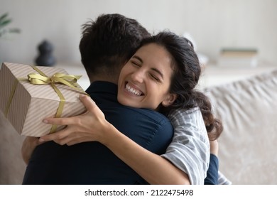Grateful happy young woman holding festive wrap, hugging beloved husband, boyfriend, thanking for present. Sweet millennial couple celebrating anniversary, 8 march, birthday, exchanging gifts
