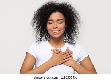 Grateful hopeful happy black woman holding hands on chest feeling pleased thankful, sincere african lady expressing heartfelt love appreciation gratitude honesty isolated on white studio background