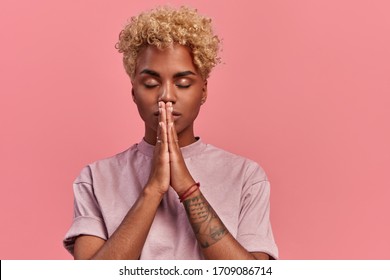 Grateful hopeful young black woman stands in meditative pose, enjoys peaceful atmosphere, holds hands in praying gesture, isolated over pink background, has sense of inner peace. Meditation concept.