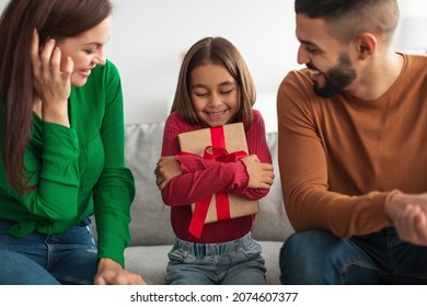 Grateful little girl sitting on couch between parents, holding and hugging wrapped gift box, man and woman greeting daughter with birthday, happy family celebrating holiday together at home