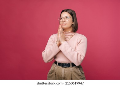 Grateful young woman is holding hands together for prayer. Studio shot over pink background.