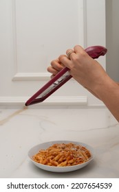 A Grater Is Held By The Hands Of A Woman, While Grating Cheese And Pouring It Onto A Plate Of Macaroni With Bolognese Sauce On A Marble Table And A Wall With Molding In The Background