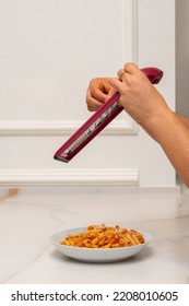 A Grater Is Held By The Hands Of A Woman, While Grating Cheese And Pouring It Onto A Plate Of Macaroni With Bolognese Sauce On A Marble Table And A Wall With Molding In The Background