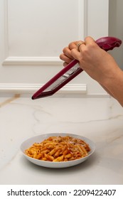 A Grater Is Held By The Hands Of A Woman, While Grating Cheese And Pouring It Onto A Plate Of Macaroni With Bolognese Sauce On A Marble Table And A Wall With Molding In The Background