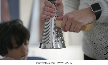 Grating cheese over a dish while wearing a smartwatch, close-up of hands holding cheese and grater, child blurred in background, home kitchen scene, preparing meal with cheese topping - Powered by Shutterstock