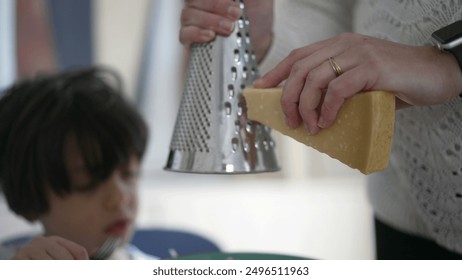 Grating cheese over a dish while wearing a smartwatch, close-up of hands holding cheese and grater, child blurred in background, home kitchen scene, preparing meal with cheese topping - Powered by Shutterstock