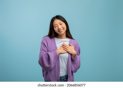 Gratitude concept. Thankful asian lady keeping both hands on chest and smiling with closed eyes, expressing appreciation and kindness while standing over blue studio background